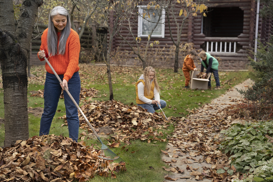 Familie sammelt gemeinsam Herbstlaub im Garten vor einem Holzhaus, Erwachsene und Kinder helfen bei der Gartenarbeit.
